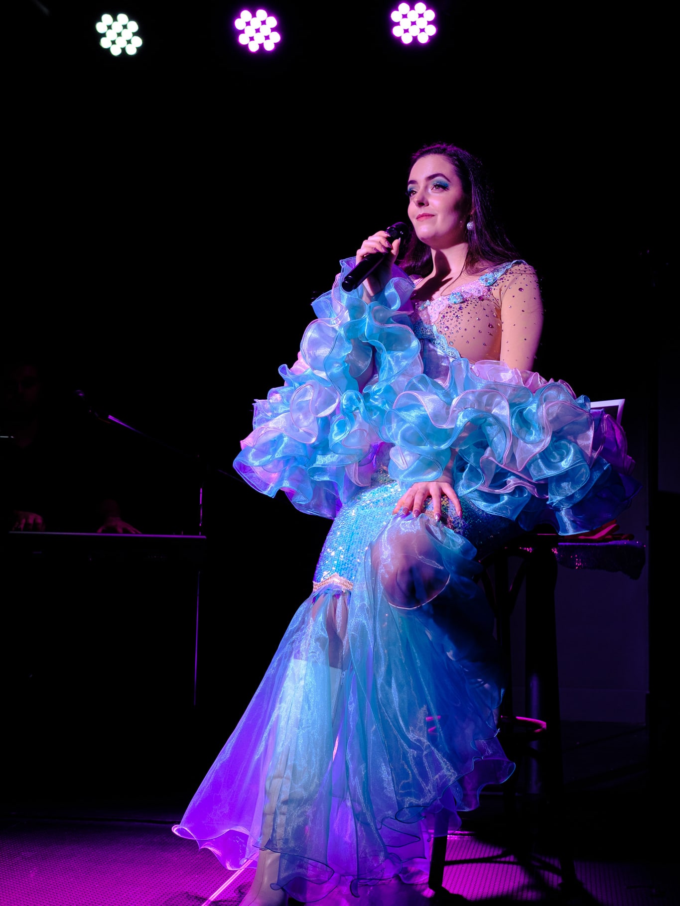 Abbey singing while seated on a stool in her ruffled costume under purple stage light