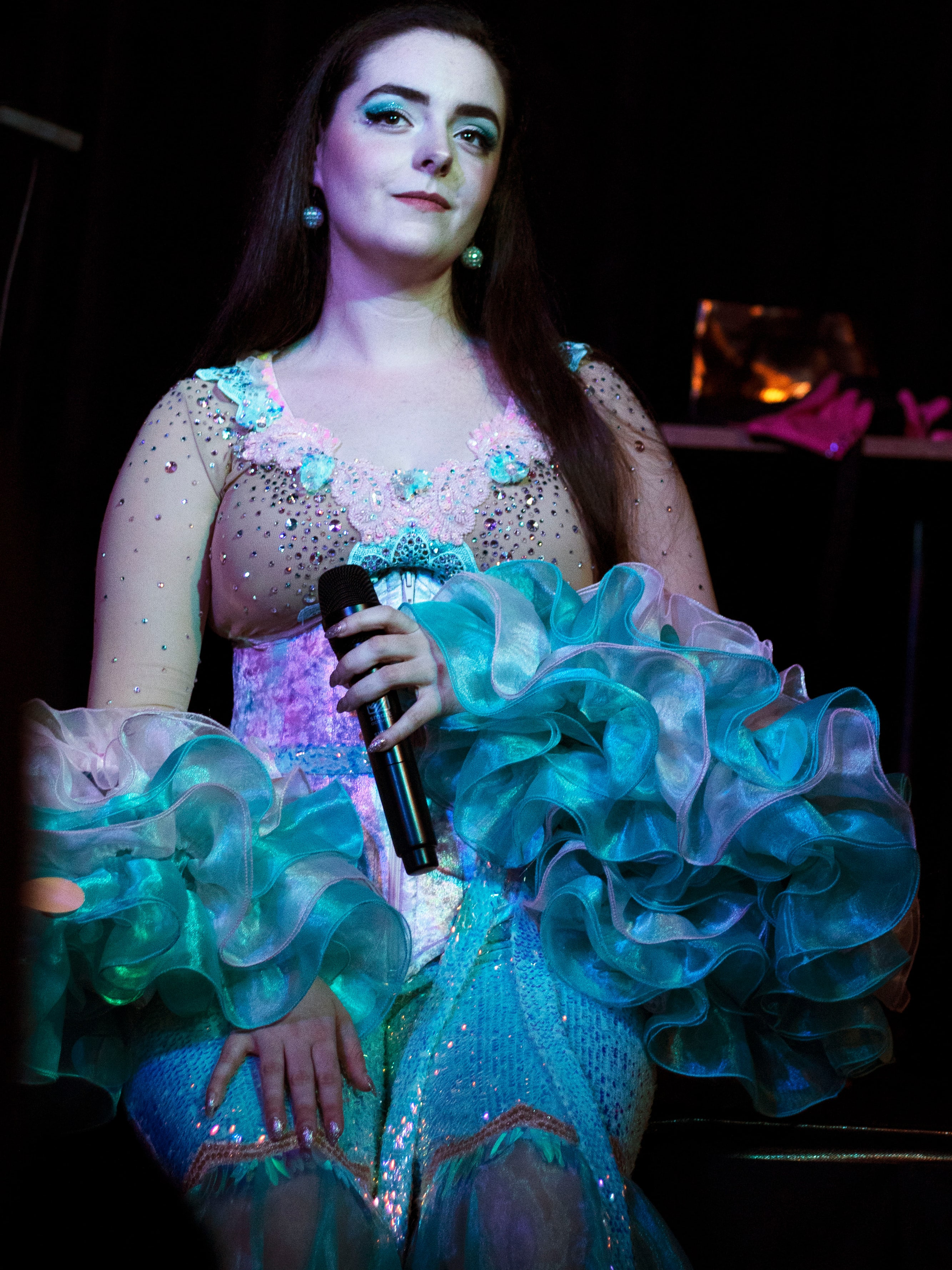 Abbey seated with a microphone in her lap, wearing a jewelled bodice and aqua ruffles