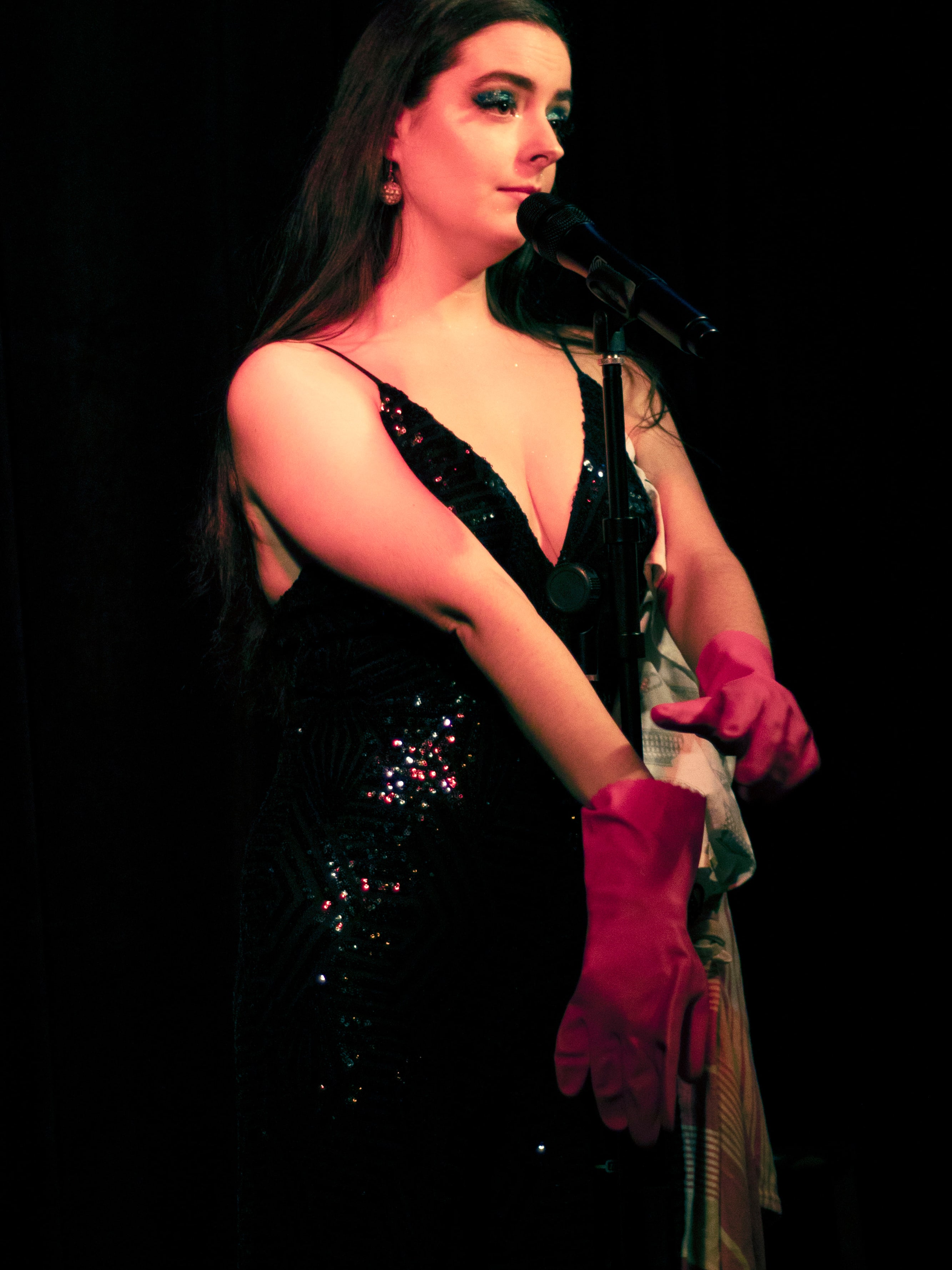 Abbey in a black sequin gown holding pink rubber gloves on stage