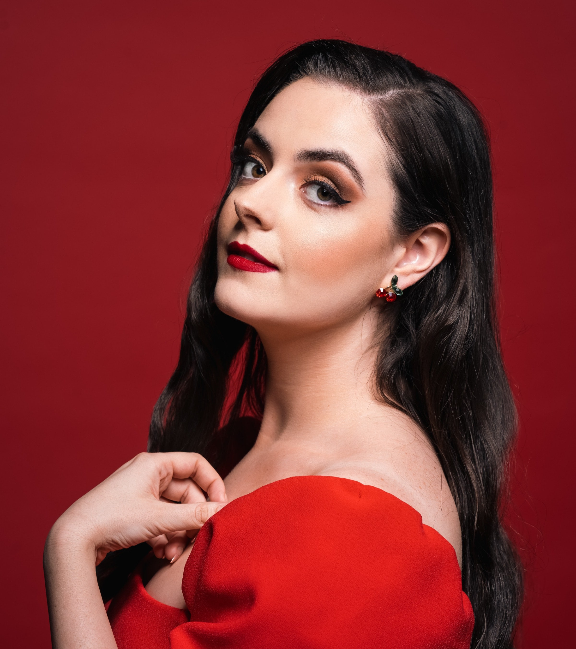 Abbey Paige Williams wears a red dress in front of a red backdrop in this headshot. Red lipstick and cherry earrings.
