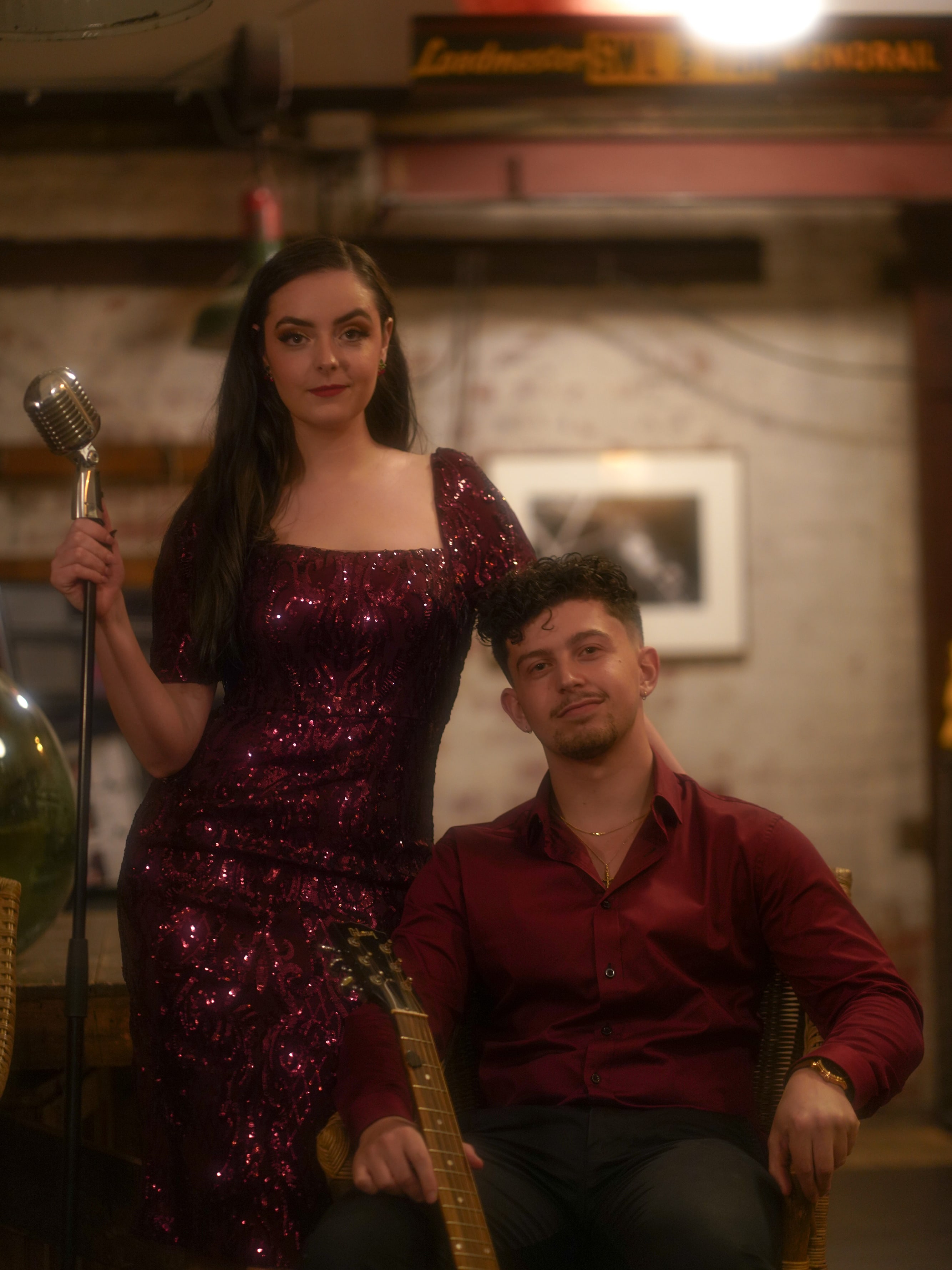 Abbey in a burgundy sequin gown standing at a vintage microphone beside her seated guitarist