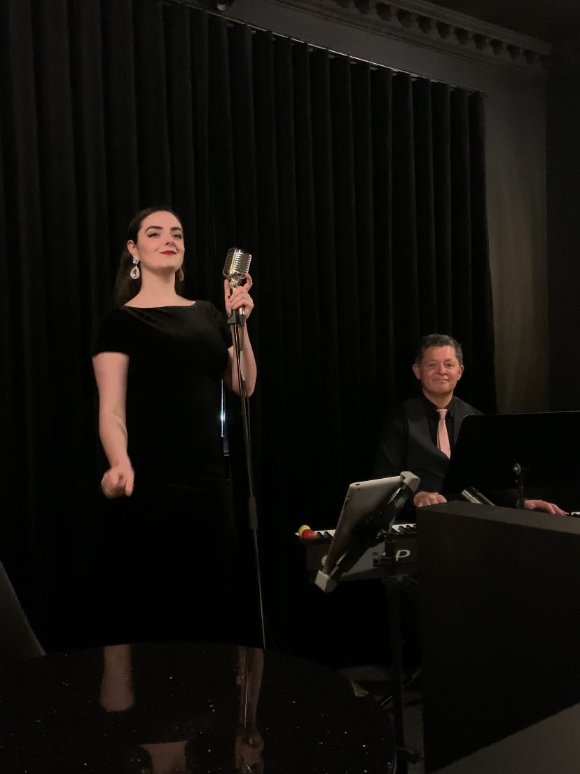 Abbey in a black dress singing at a vintage microphone while a pianist plays on a dark stage