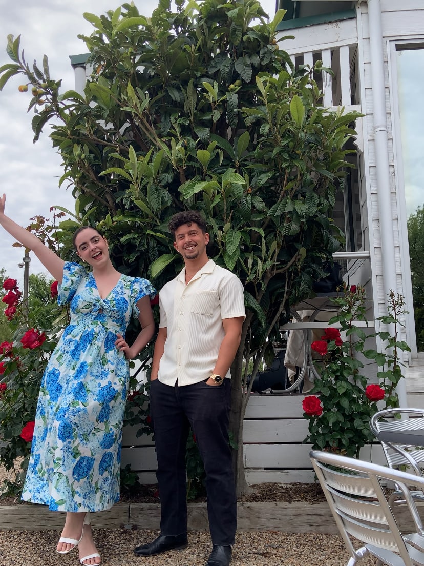 Abbey in a blue floral dress with her guitarist in a garden of red roses outside a white weatherboard building