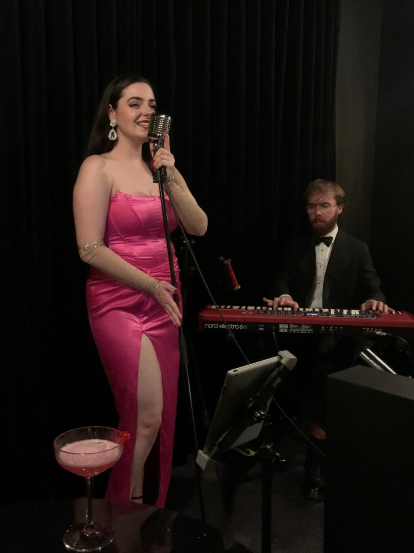 Abbey in a pink satin gown singing at a vintage microphone with a keyboard player behind her and a cocktail on a table