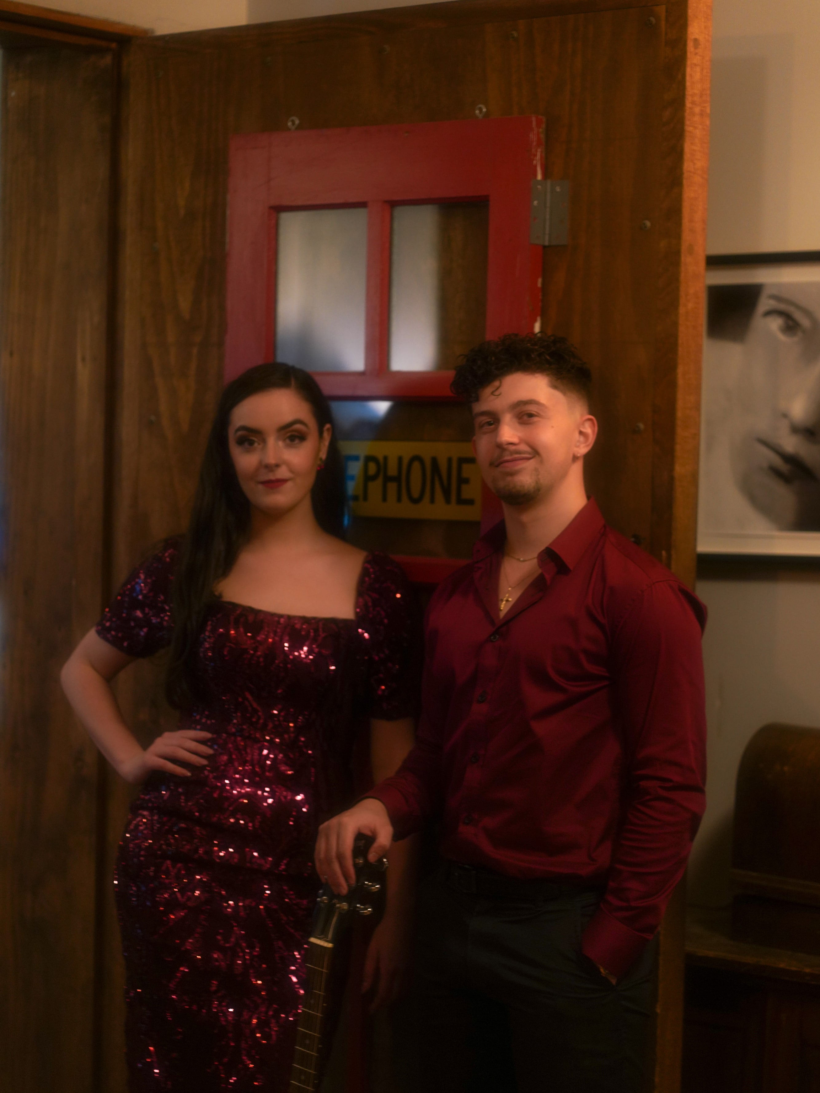 Abbey in a burgundy sequin gown and her guitarist in a red shirt standing by a vintage telephone booth