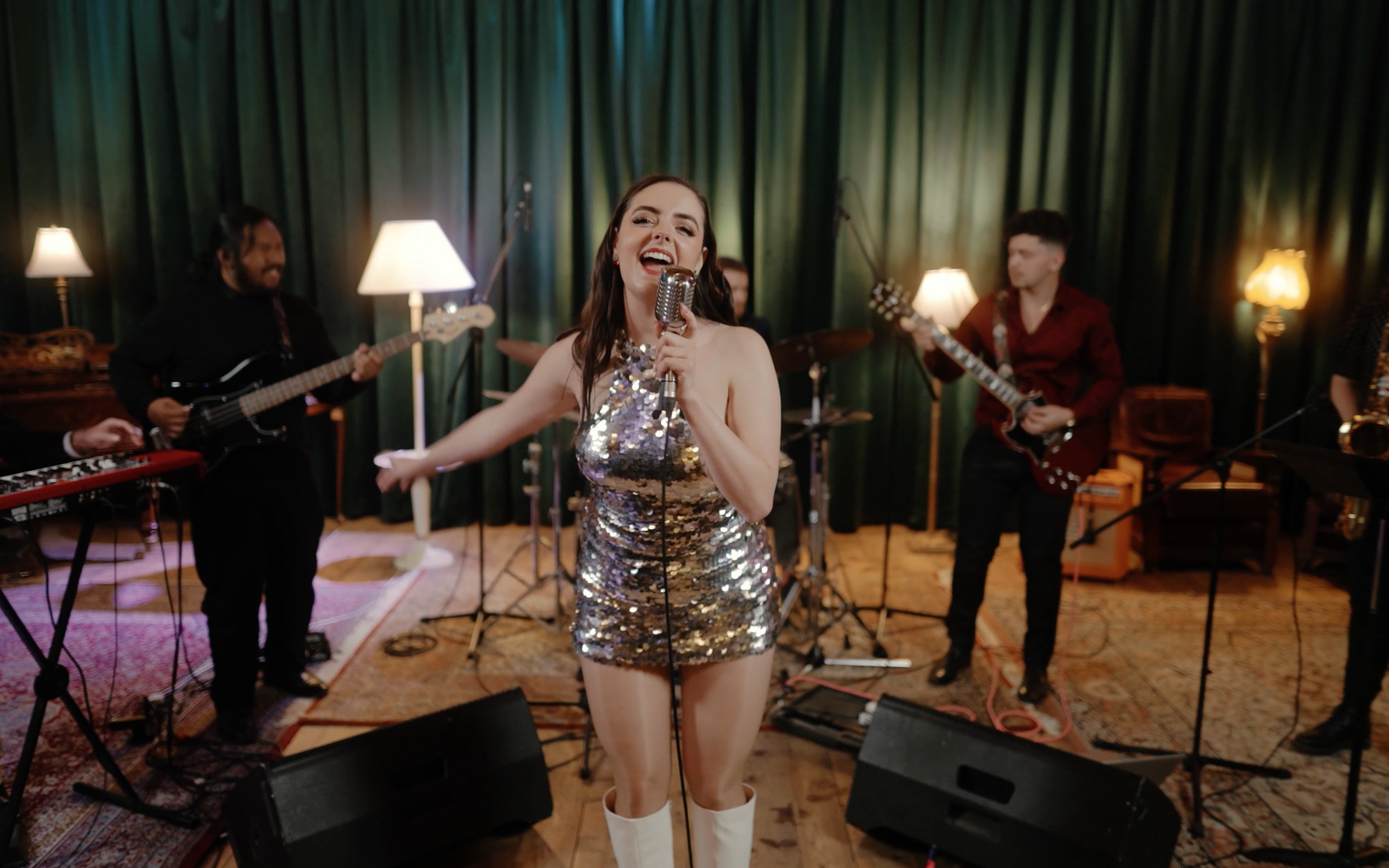 Abbey singing in a silver sequin playsuit and white boots, with Cherry Lane playing behind her in front of green velvet curtains
