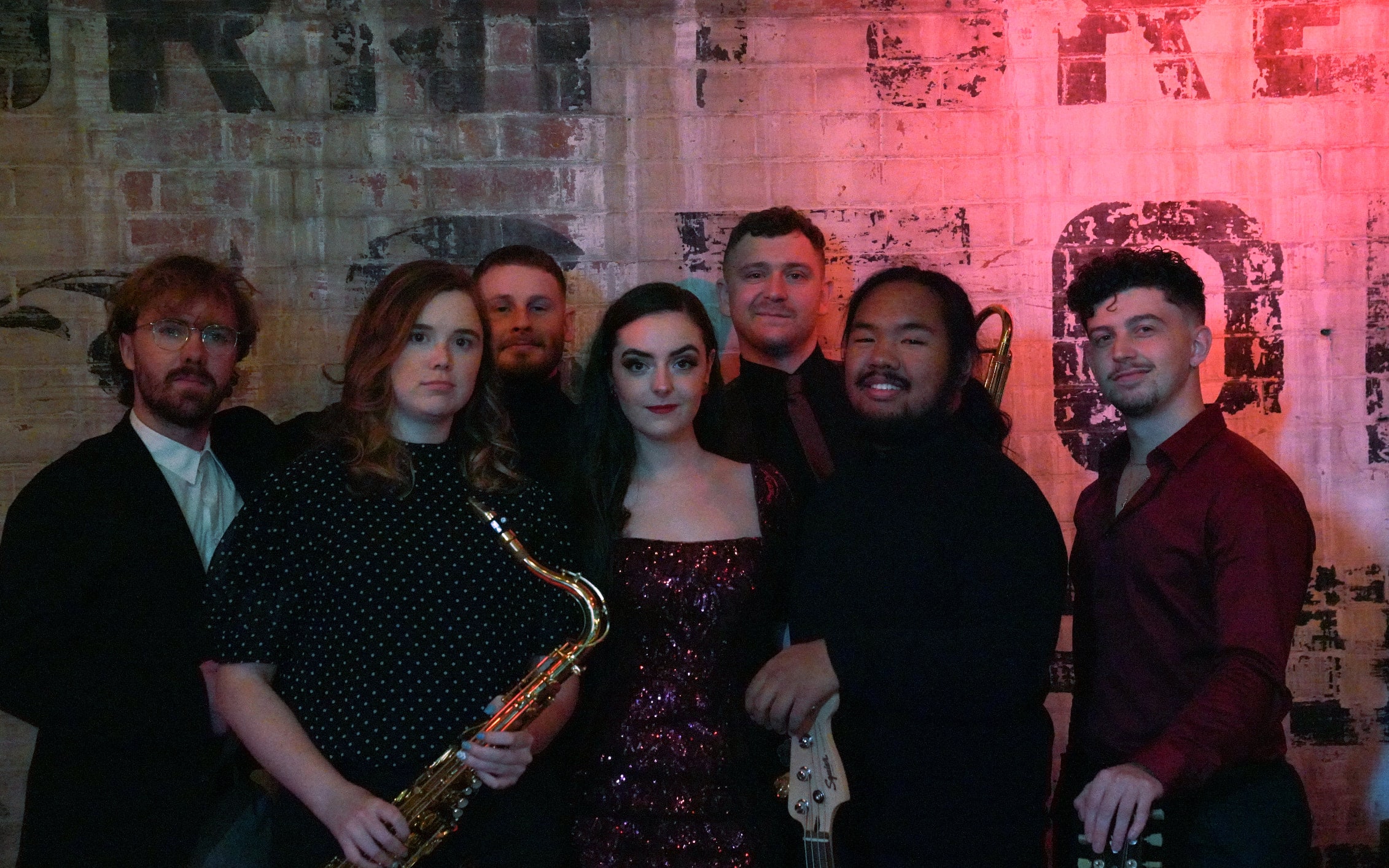 The Cherry Lane band posing together in front of a graffiti-covered brick wall, the saxophonist holding her saxophone
