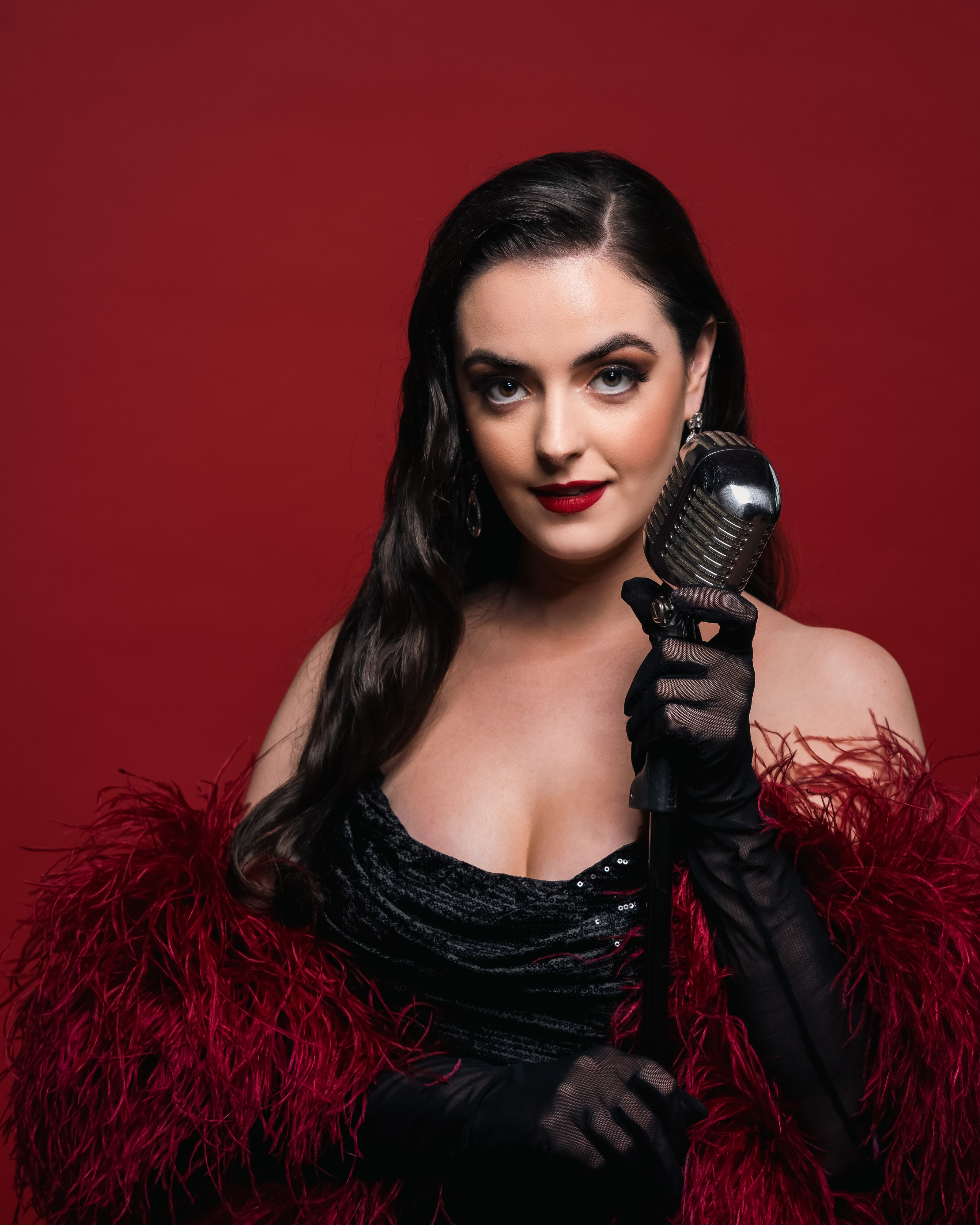 Abbey in a black gown and red feather stole holding a vintage microphone against a red backdrop