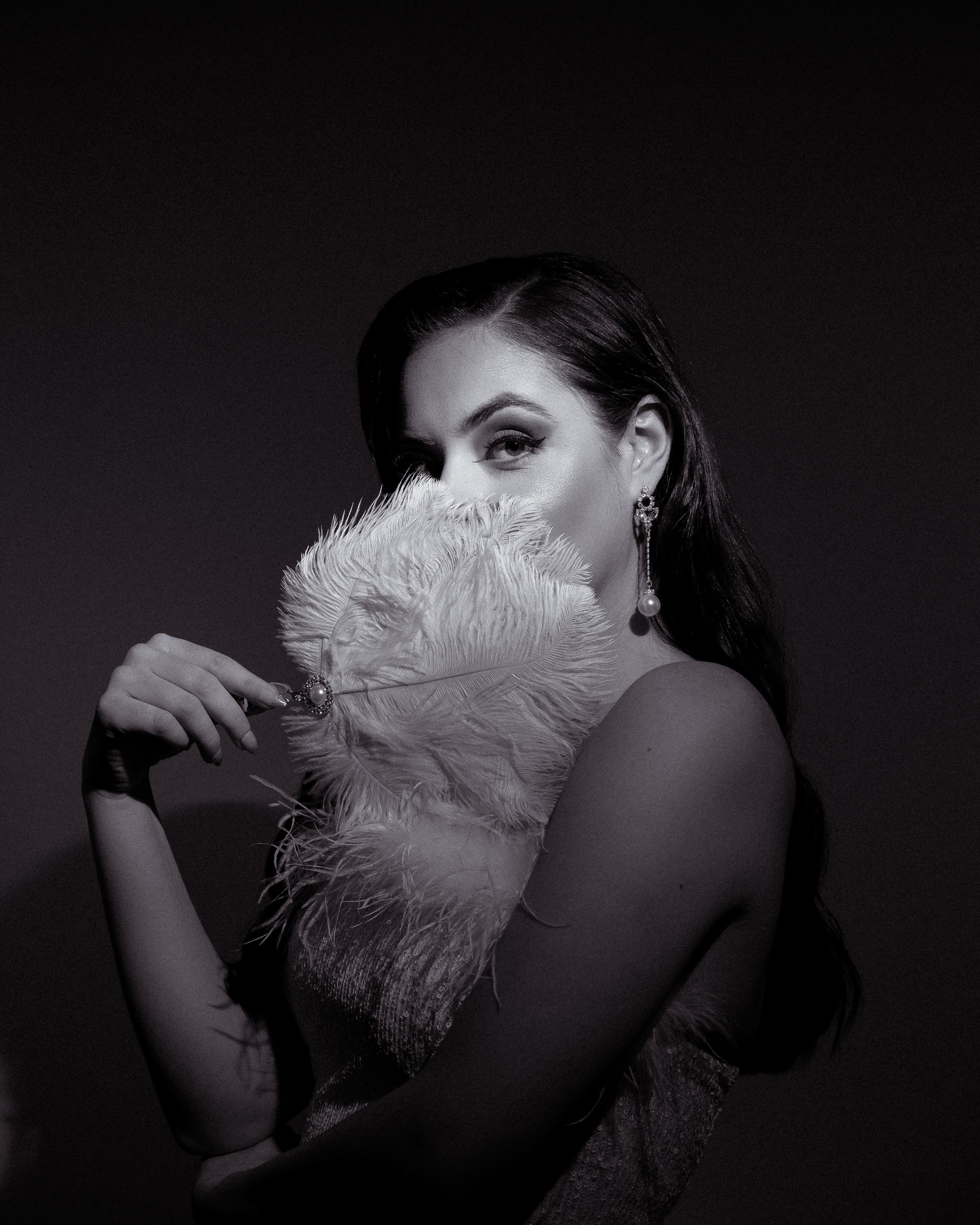 Black and white portrait of Abbey Paige Williams holding a white feather fan over the lower half of her face, wearing pearl drop earrings