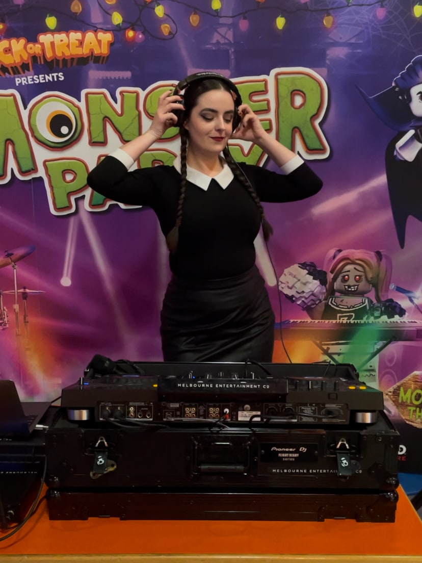 Abbey DJing in a black dress with a white collar, wearing headphones, in front of a Monster Party banner