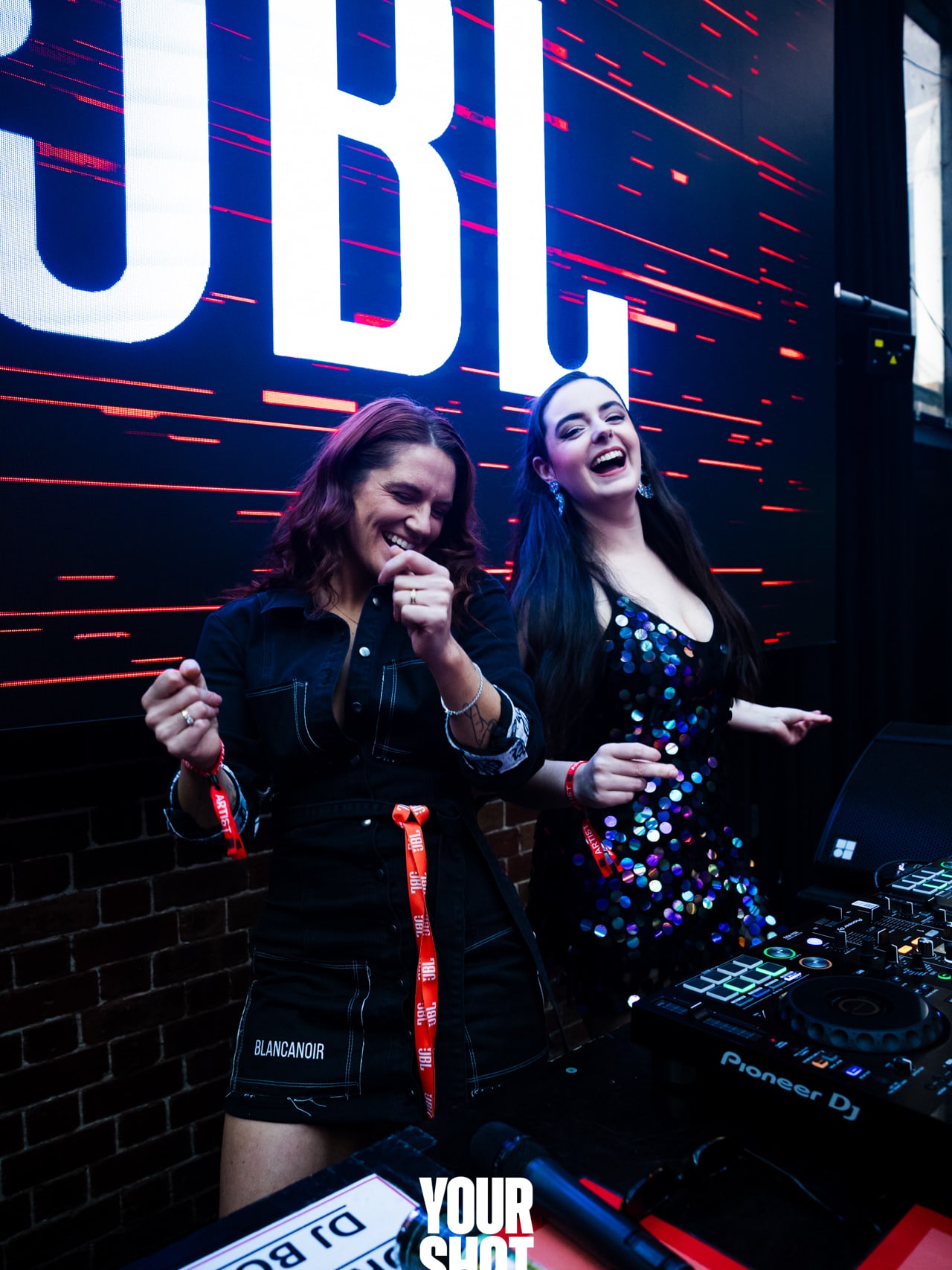 Abbey and Cass laughing together in the DJ booth under a JBL sign at the YOURSHOT DJ competition