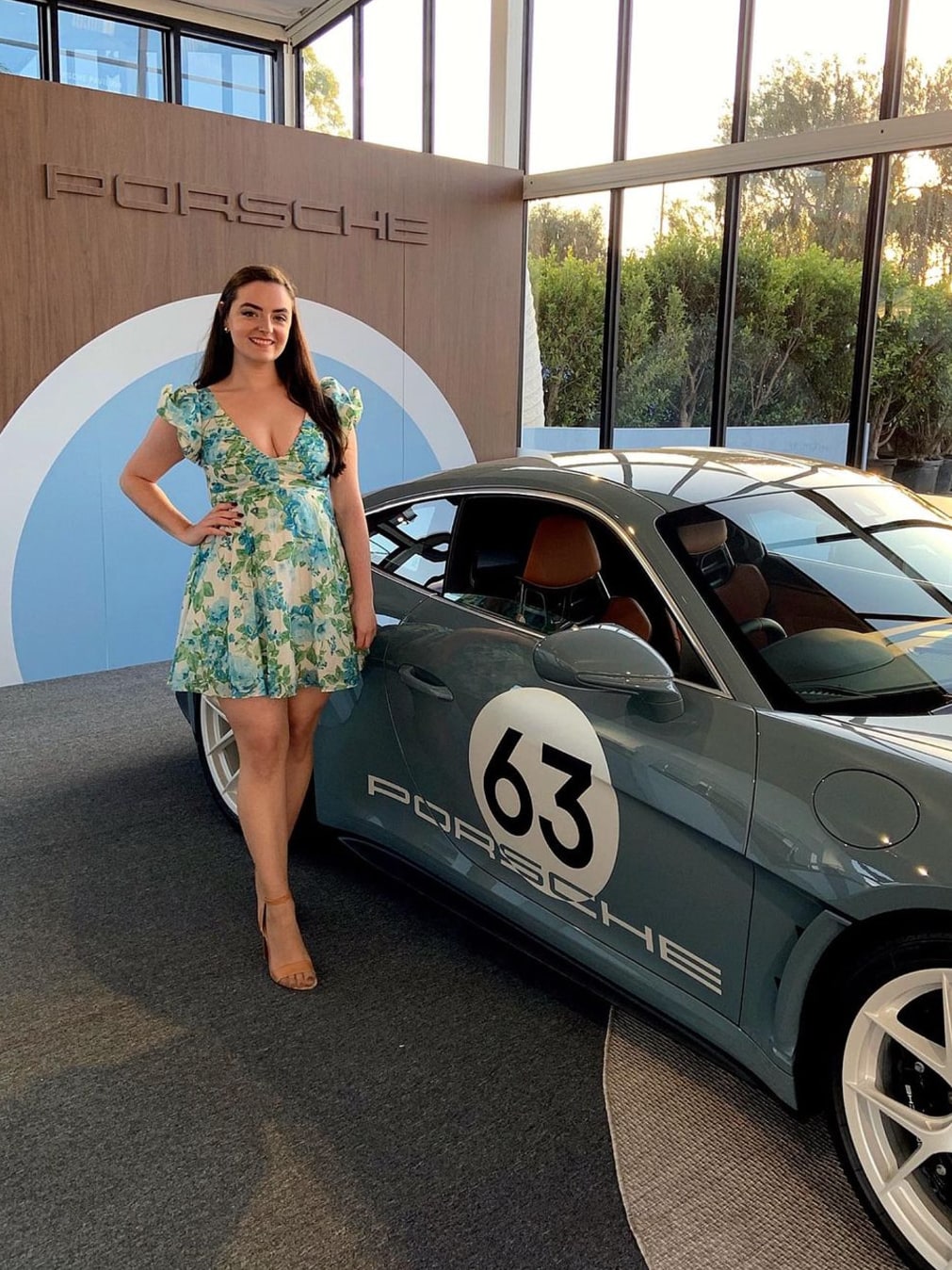 Abbey in a floral mini dress beside a blue Porsche race car in the Porsche Pavilion
