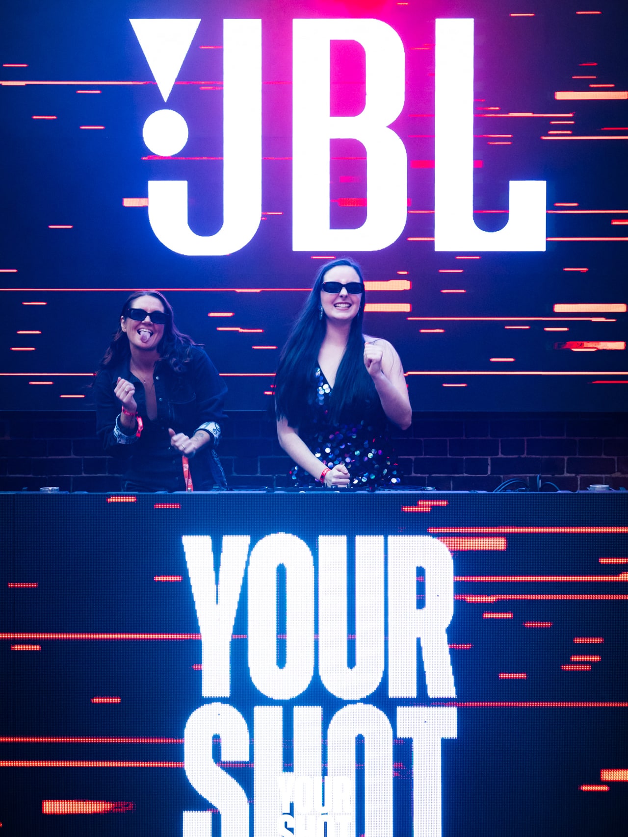Abbey and Cass in sunglasses DJing beneath the JBL and YOURSHOT screens