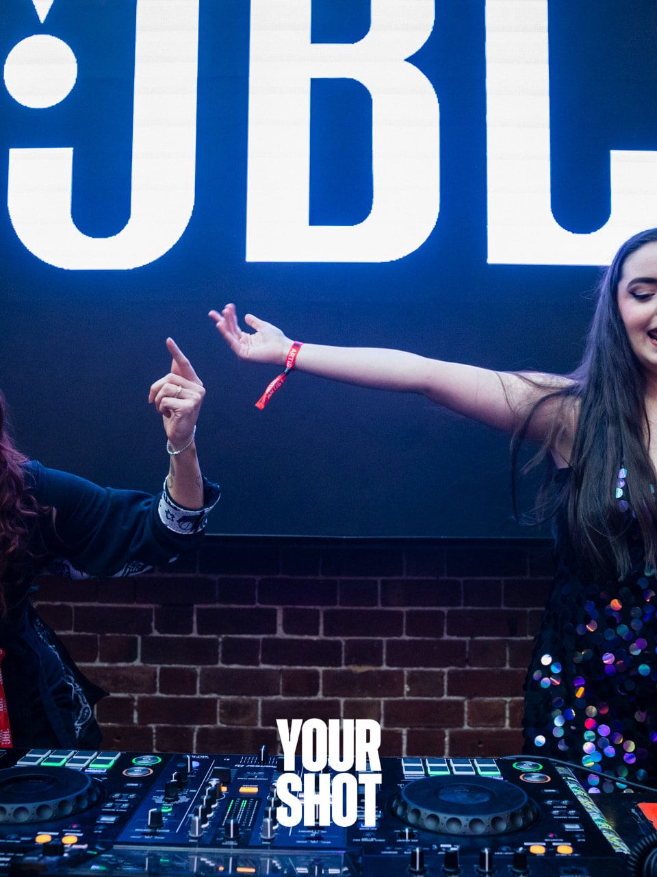 Abbey and Cass pointing and dancing behind the decks under a large JBL sign