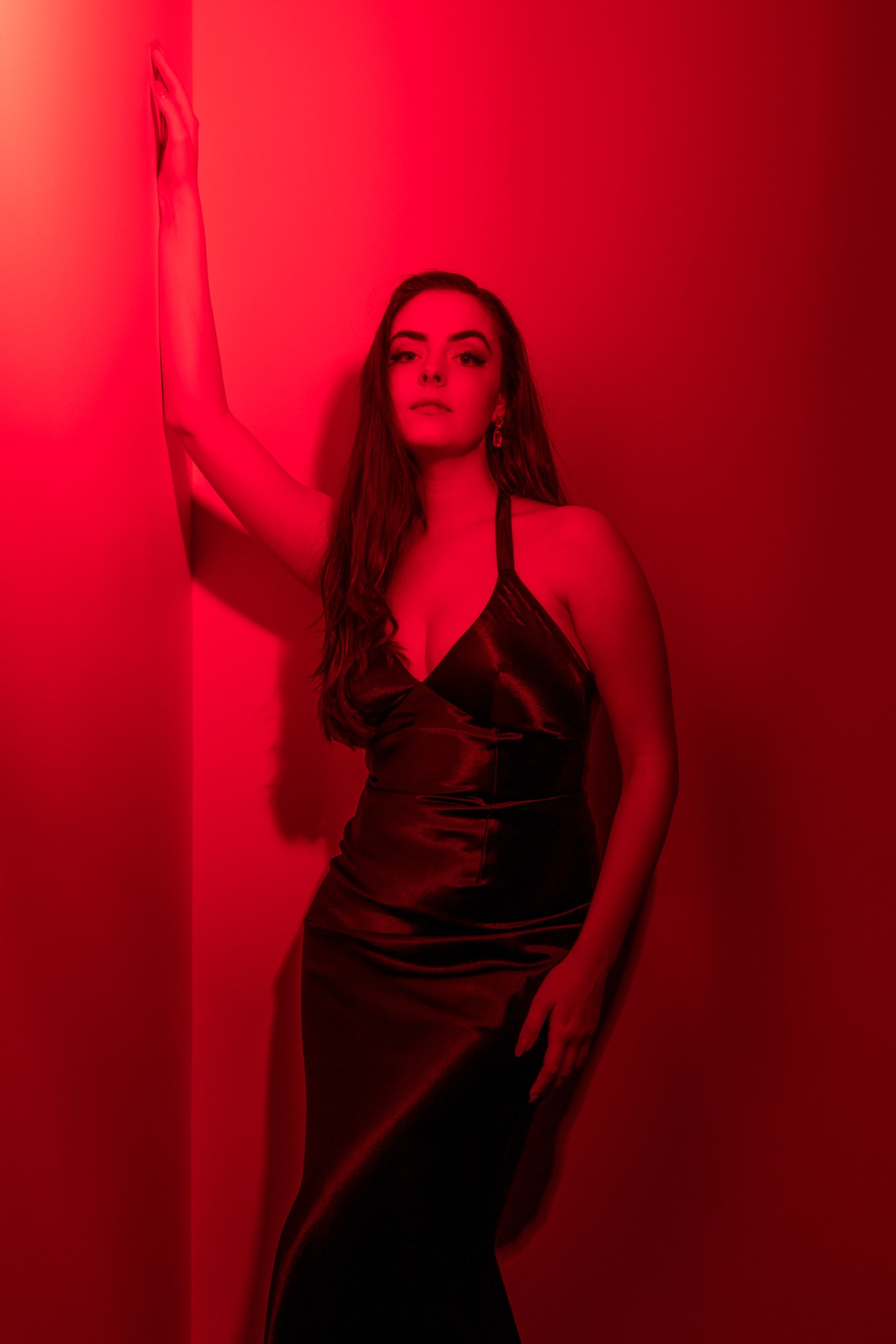 Abbey in a black satin slip dress leaning against a wall bathed in red light