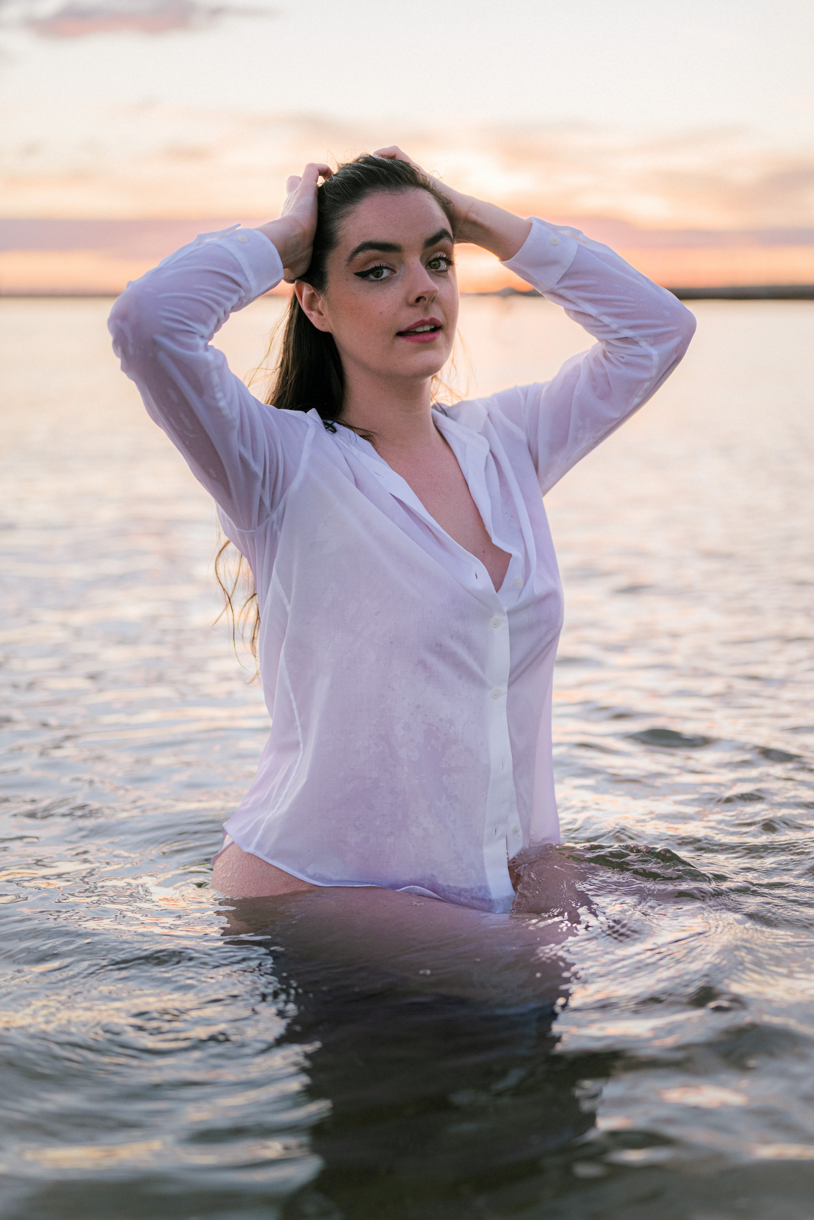 Abbey standing waist-deep in the water at sunset in a white shirt, hands in her hair