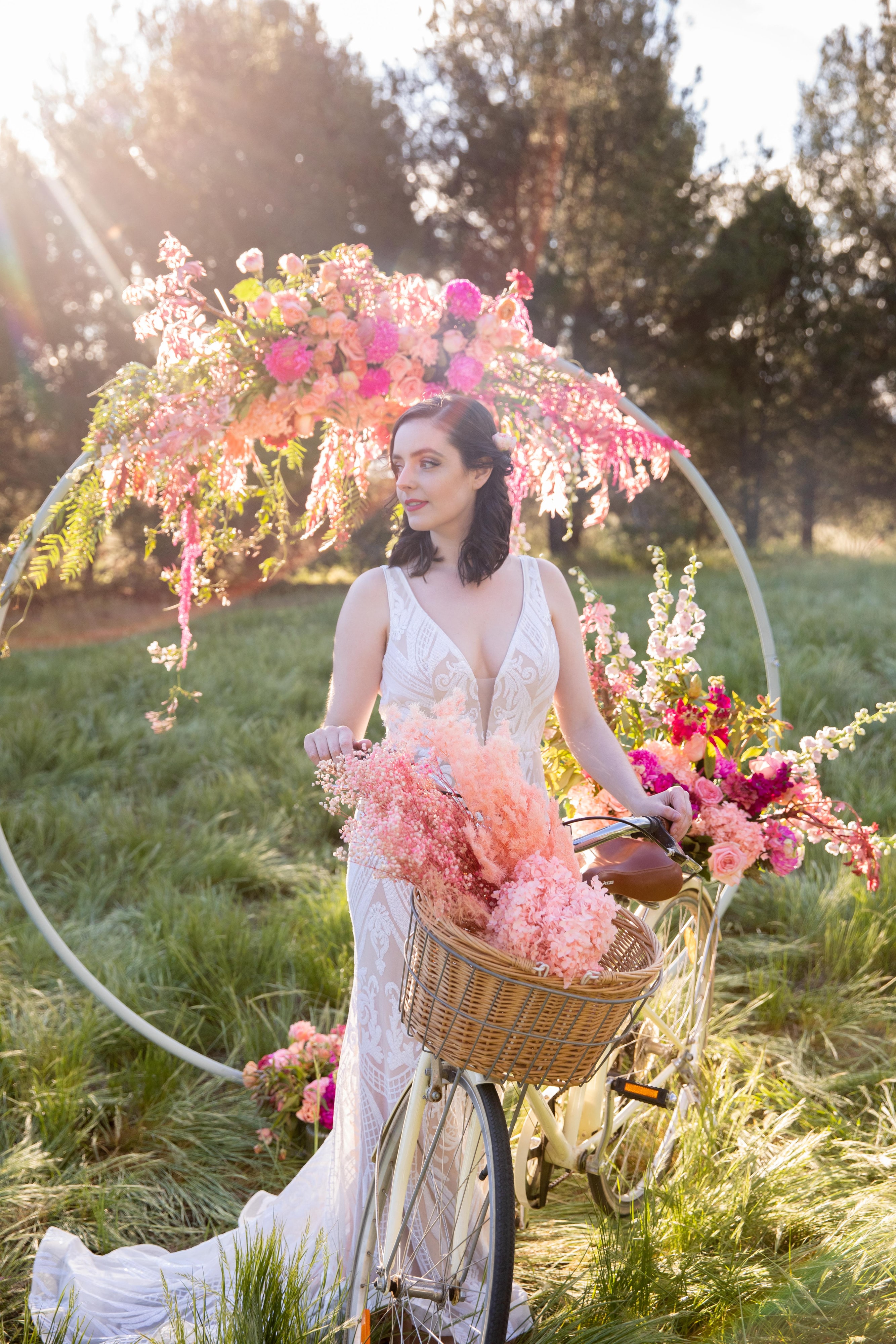 Abbey in a white bridal gown holding a bicycle with a basket of pink flowers beneath a floral arch