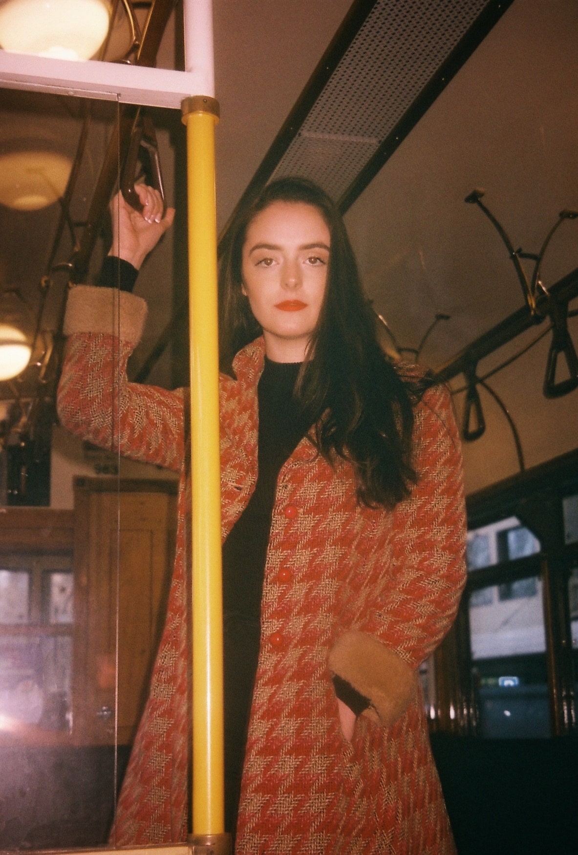 Abbey in a red houndstooth coat holding a pole inside a vintage tram