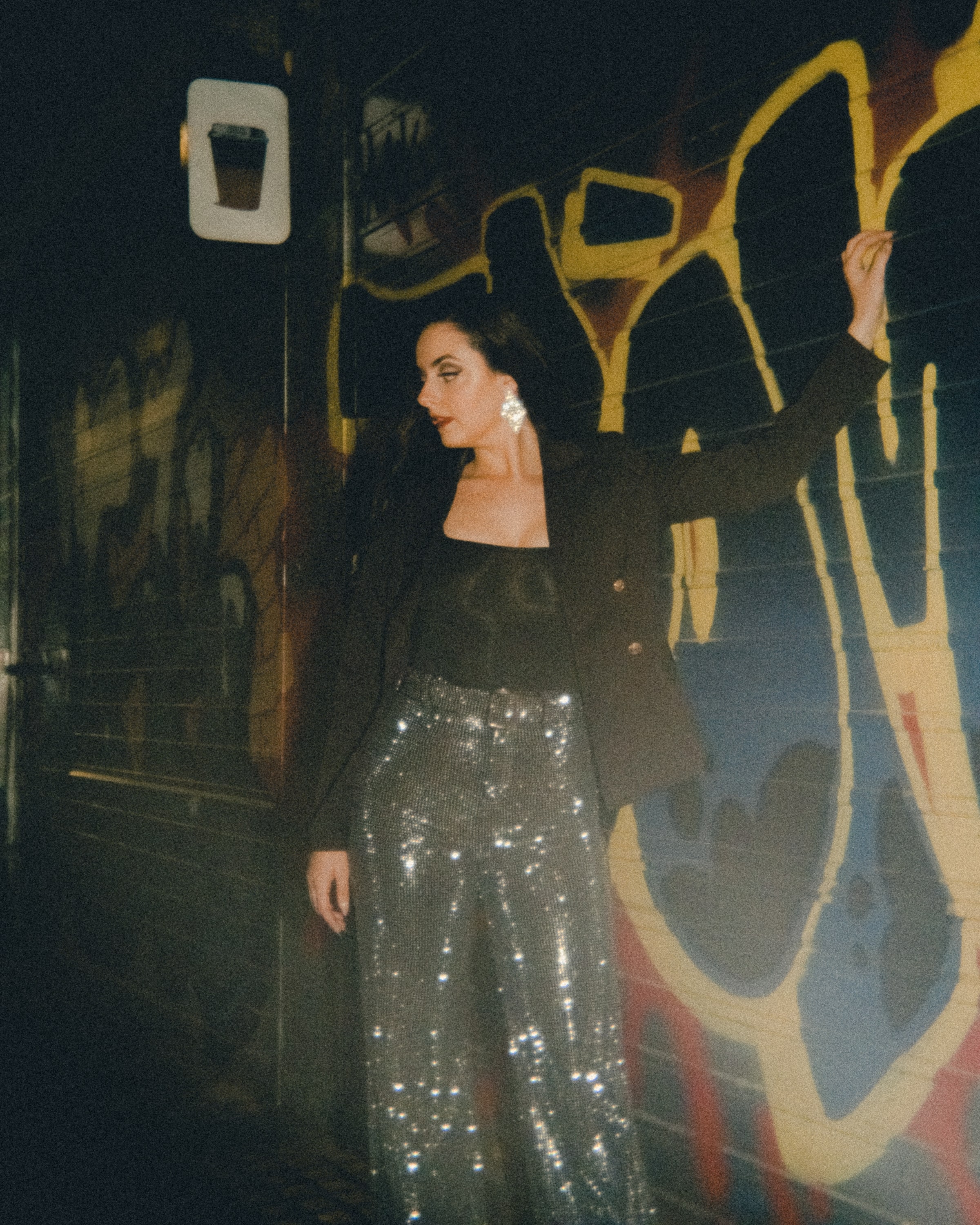Abbey in sequin trousers and a black blazer leaning against a graffiti wall at night