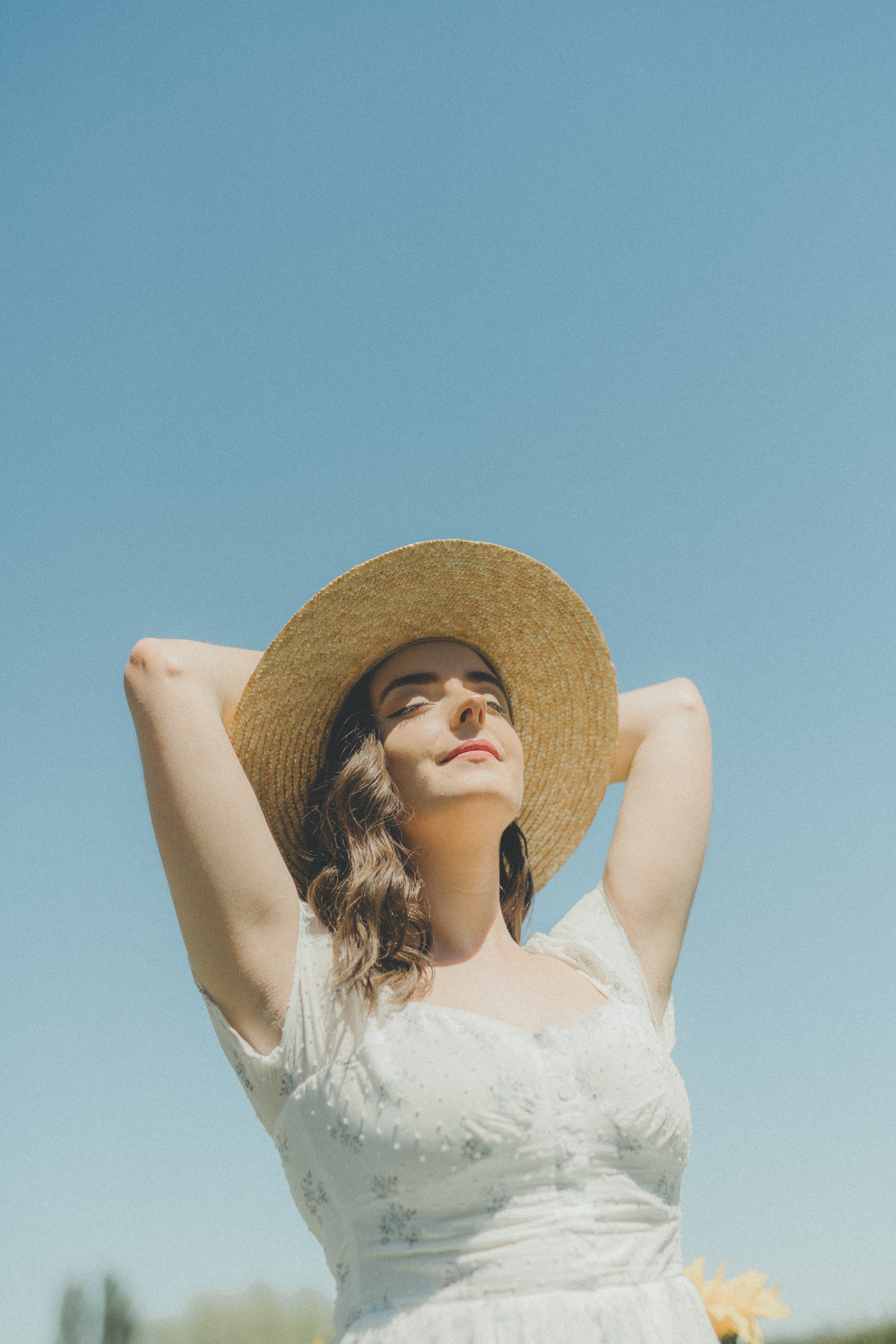 Abbey in a straw hat and white dress with her arms raised against a blue sky