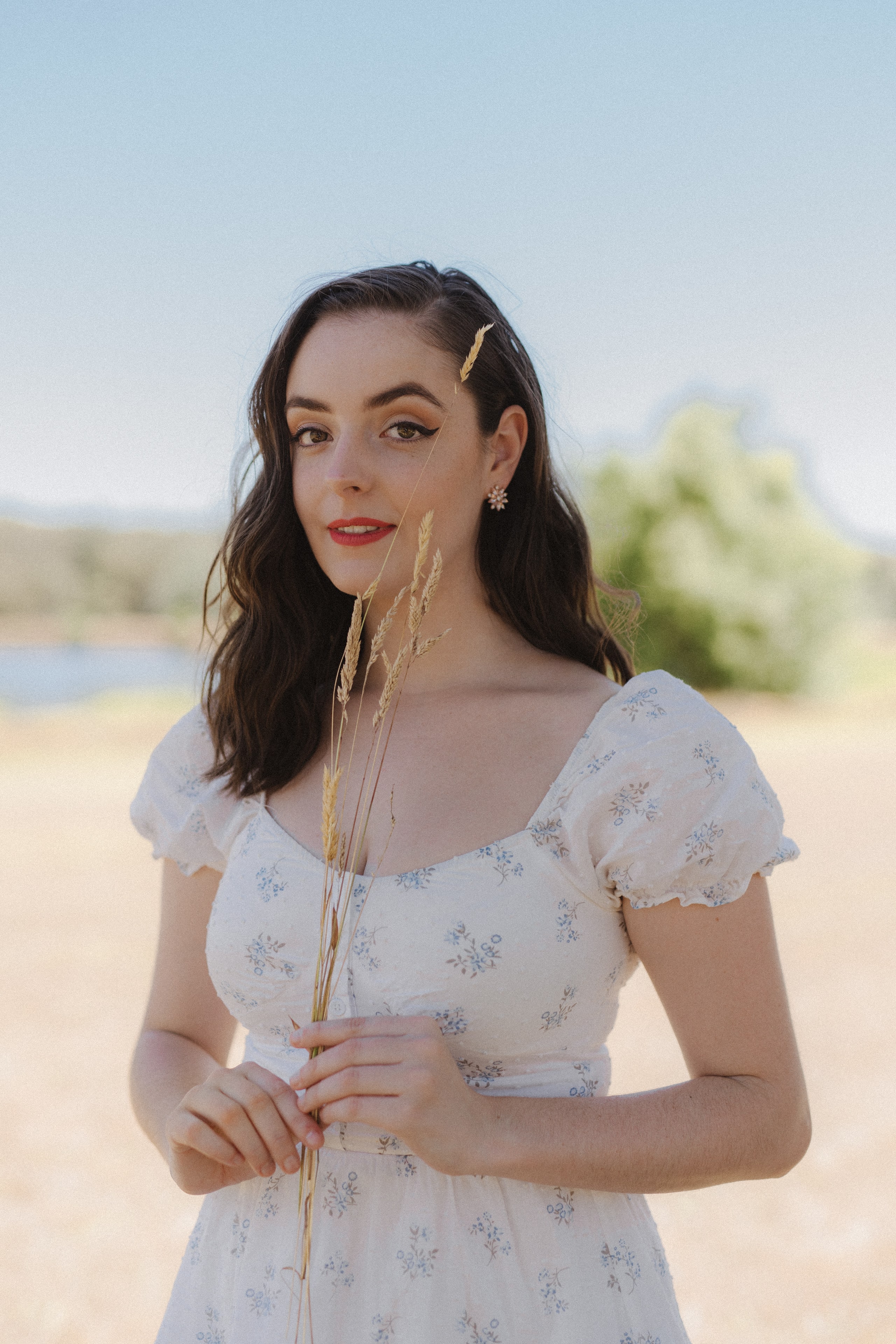 Abbey in a white floral puff-sleeve dress holding dried grass by a lake