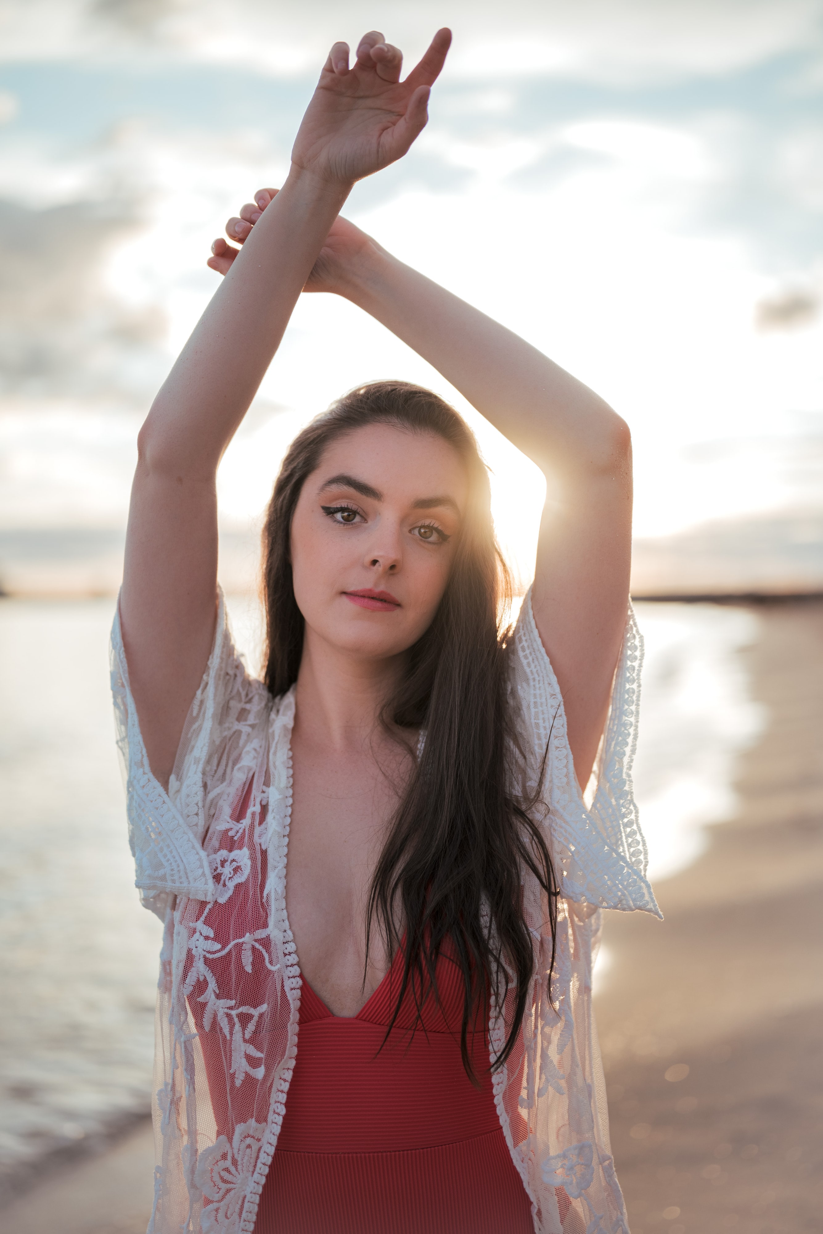 Abbey in a red top and sheer lace kimono with her arms raised above her head at the beach