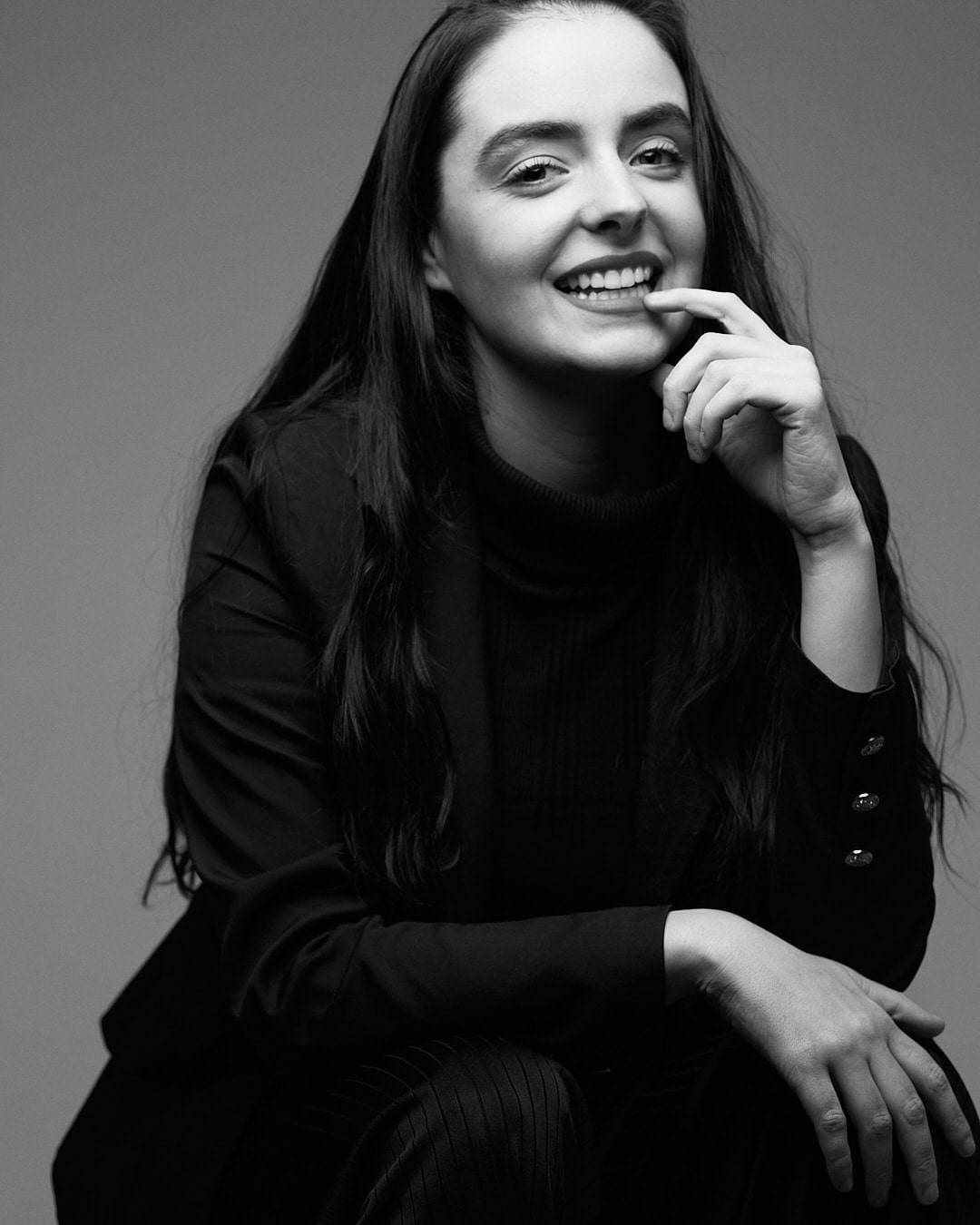 Black and white portrait of Abbey smiling with her finger to her lip, wearing a black blazer