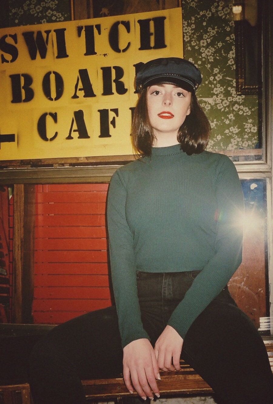 Abbey in a green jumper and baker boy cap sitting under a Switch Board Cafe sign