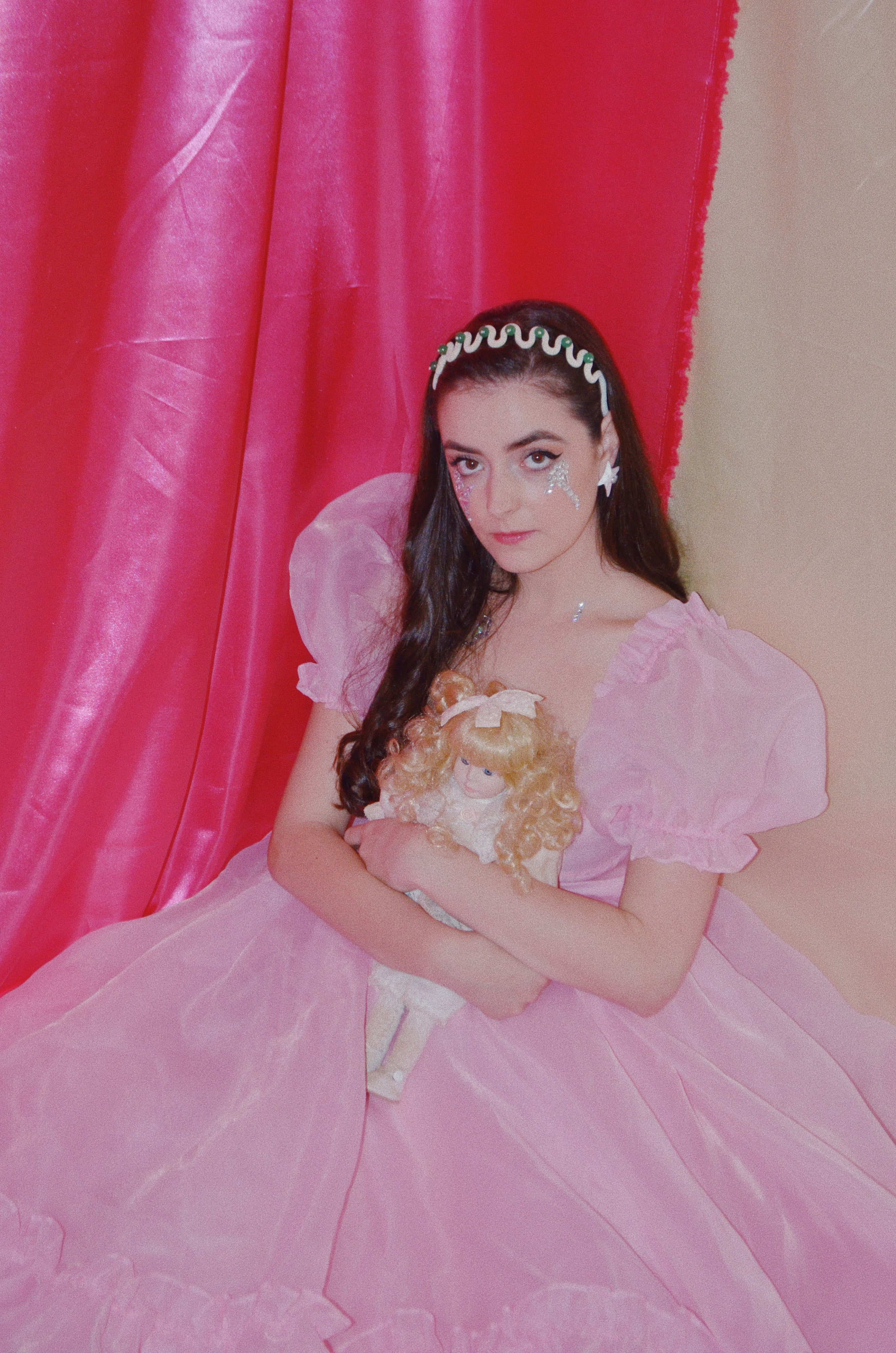 Abbey in a pink princess gown and jewelled headband holding a doll against pink drapes