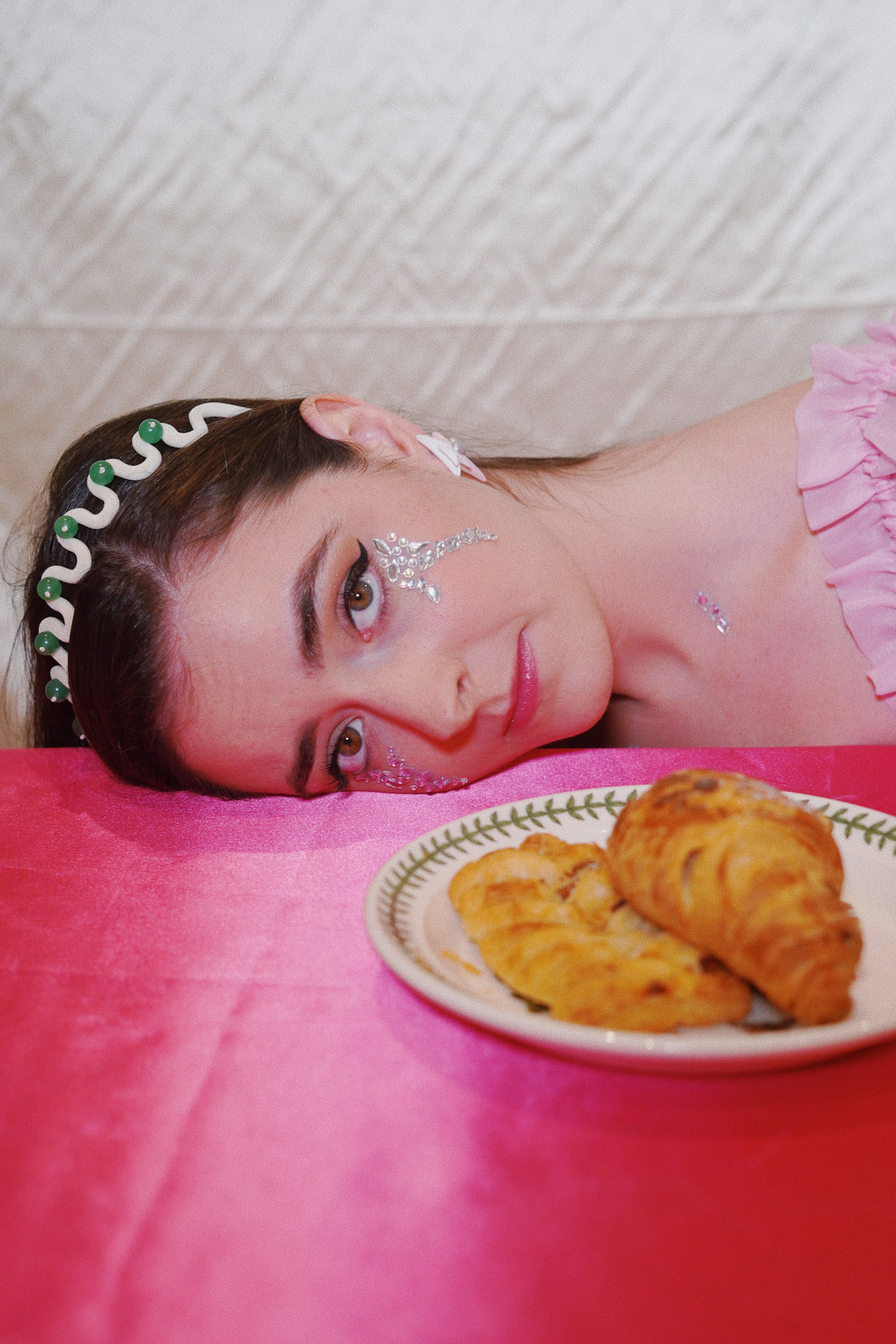 Abbey resting her head on a pink table beside a plate of croissants, wearing a jewelled headband