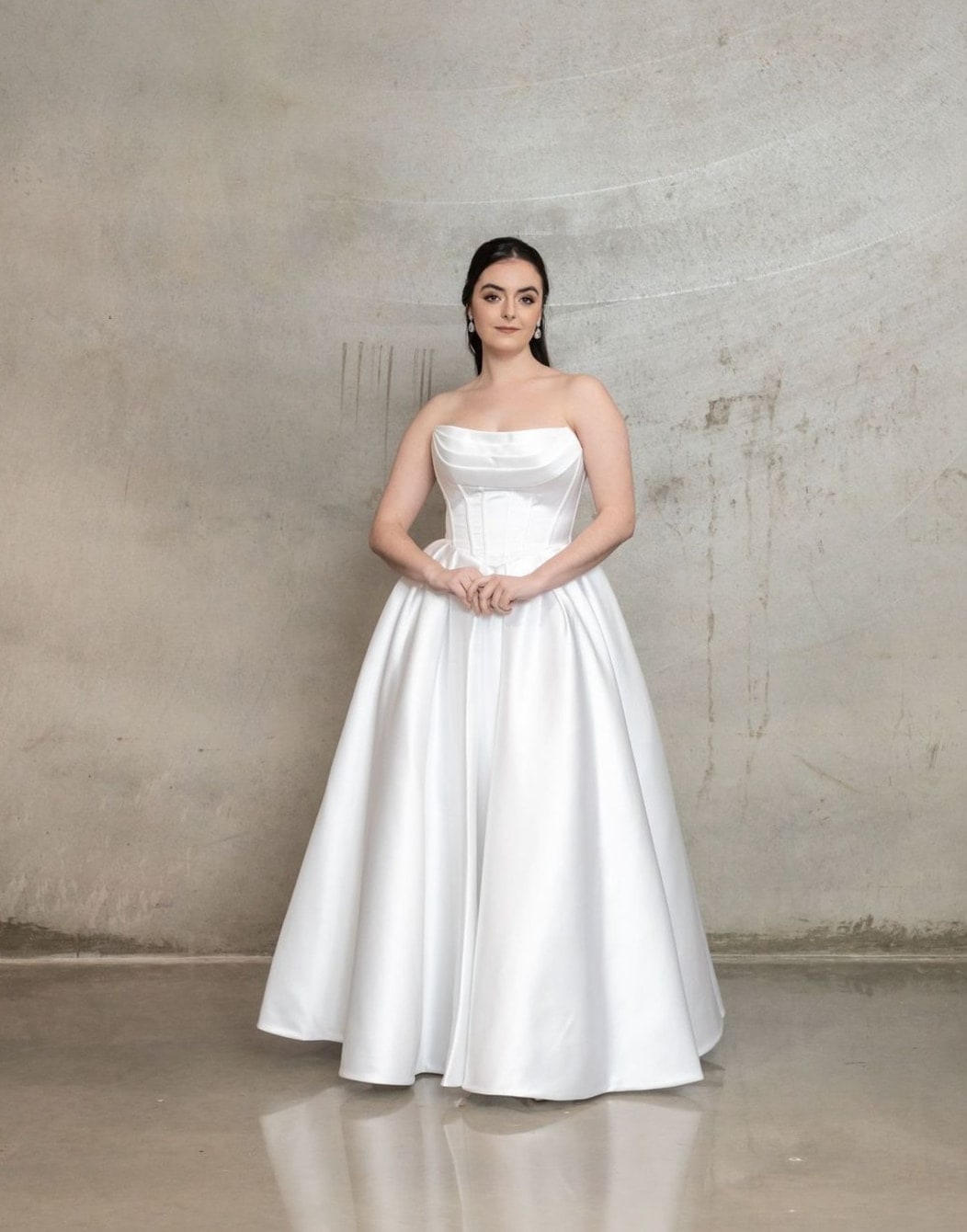 Abbey in a white strapless satin ball gown against a concrete wall