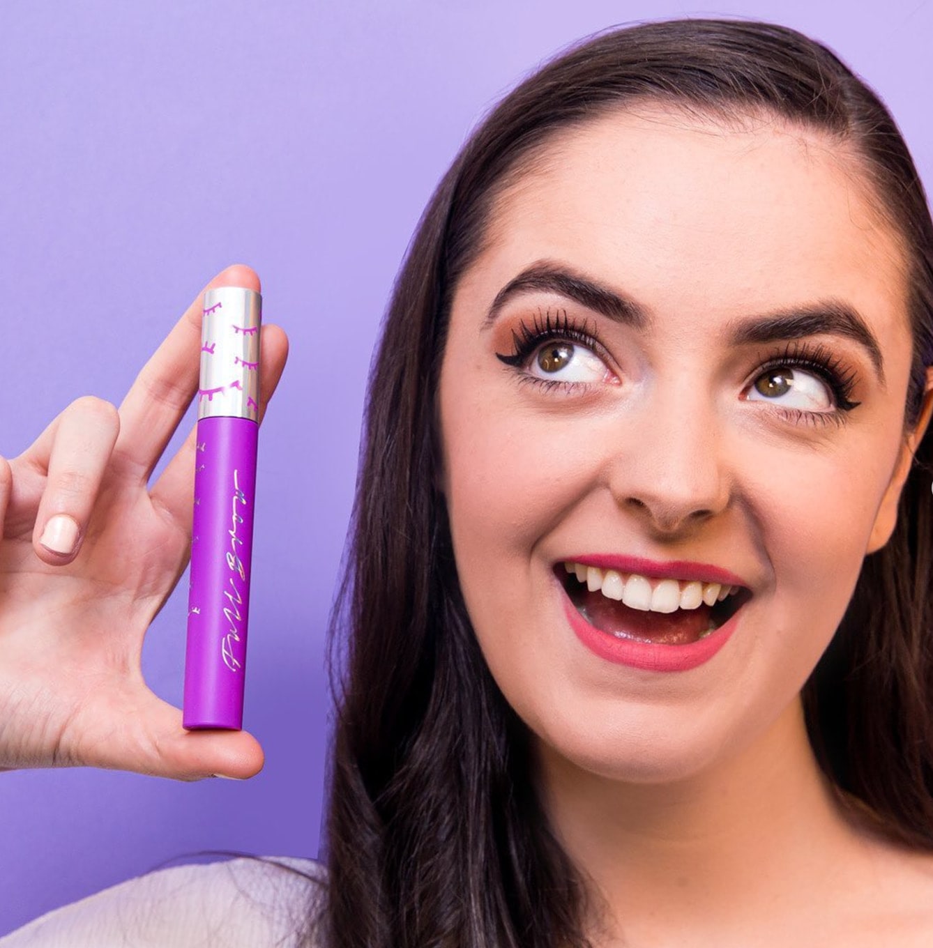 Abbey smiling and holding up a purple mascara tube against a lilac background