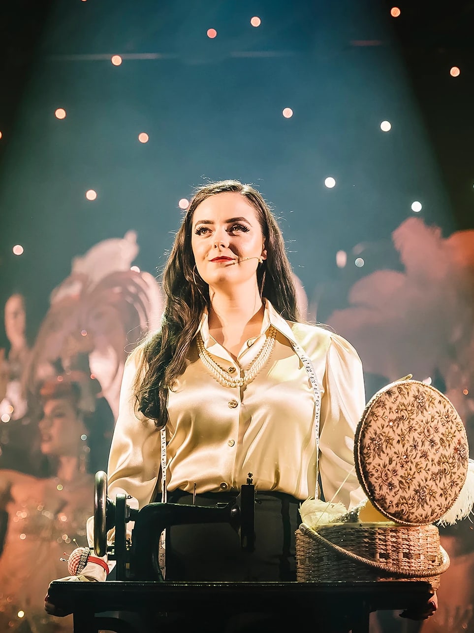 Abbey as Lady Shimmery in a satin blouse at her sewing machine on stage, dancers with feather fans behind her