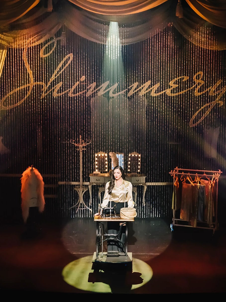Lady Shimmery seated at her sewing machine in a spotlight beneath the glittering Shimmery curtain