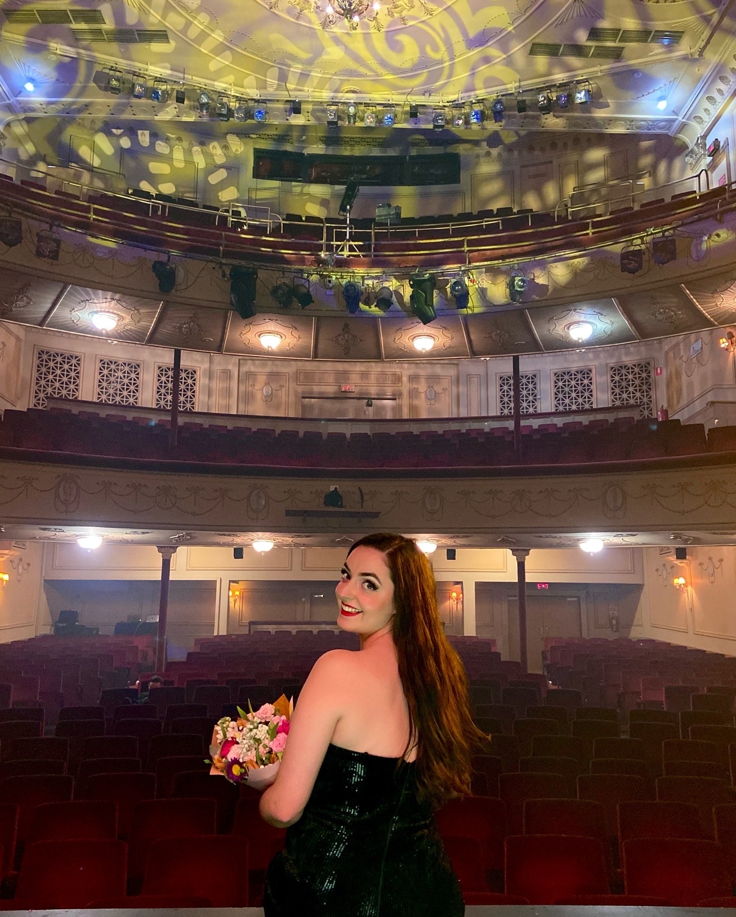 Abbey holding a bouquet on the Athenaeum Theatre stage, looking back over her shoulder at the empty auditorium