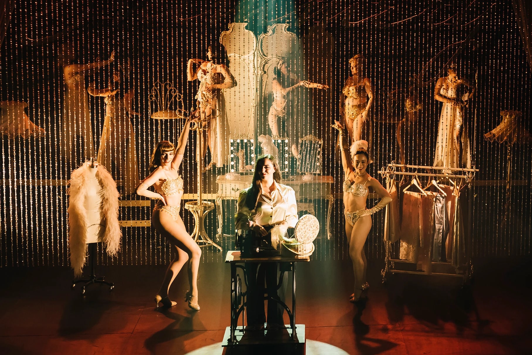 Showgirls posing on stage around Lady Shimmery at her sewing machine