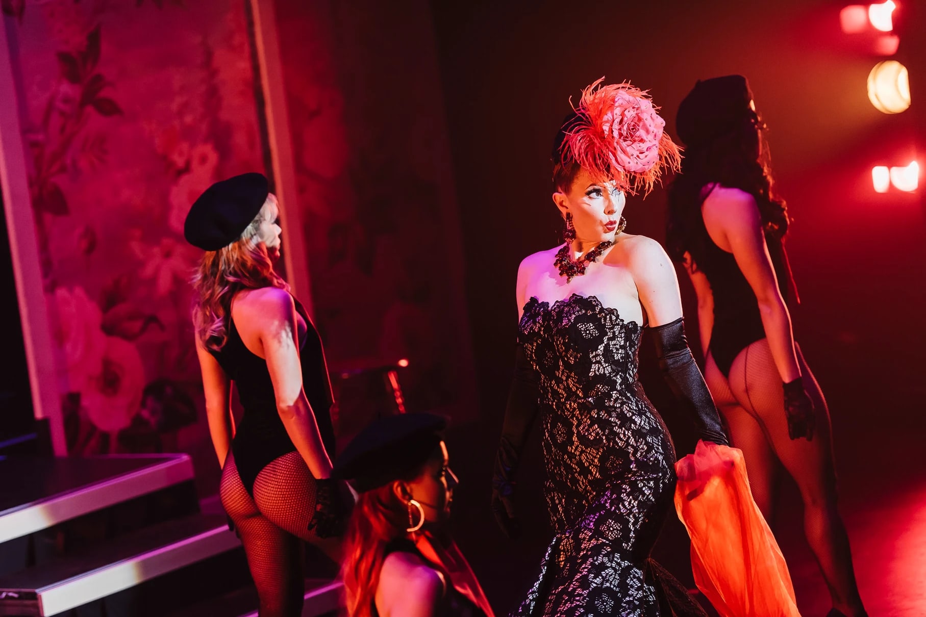 Burlesque performers in black lace and a pink feather headpiece under red stage lights