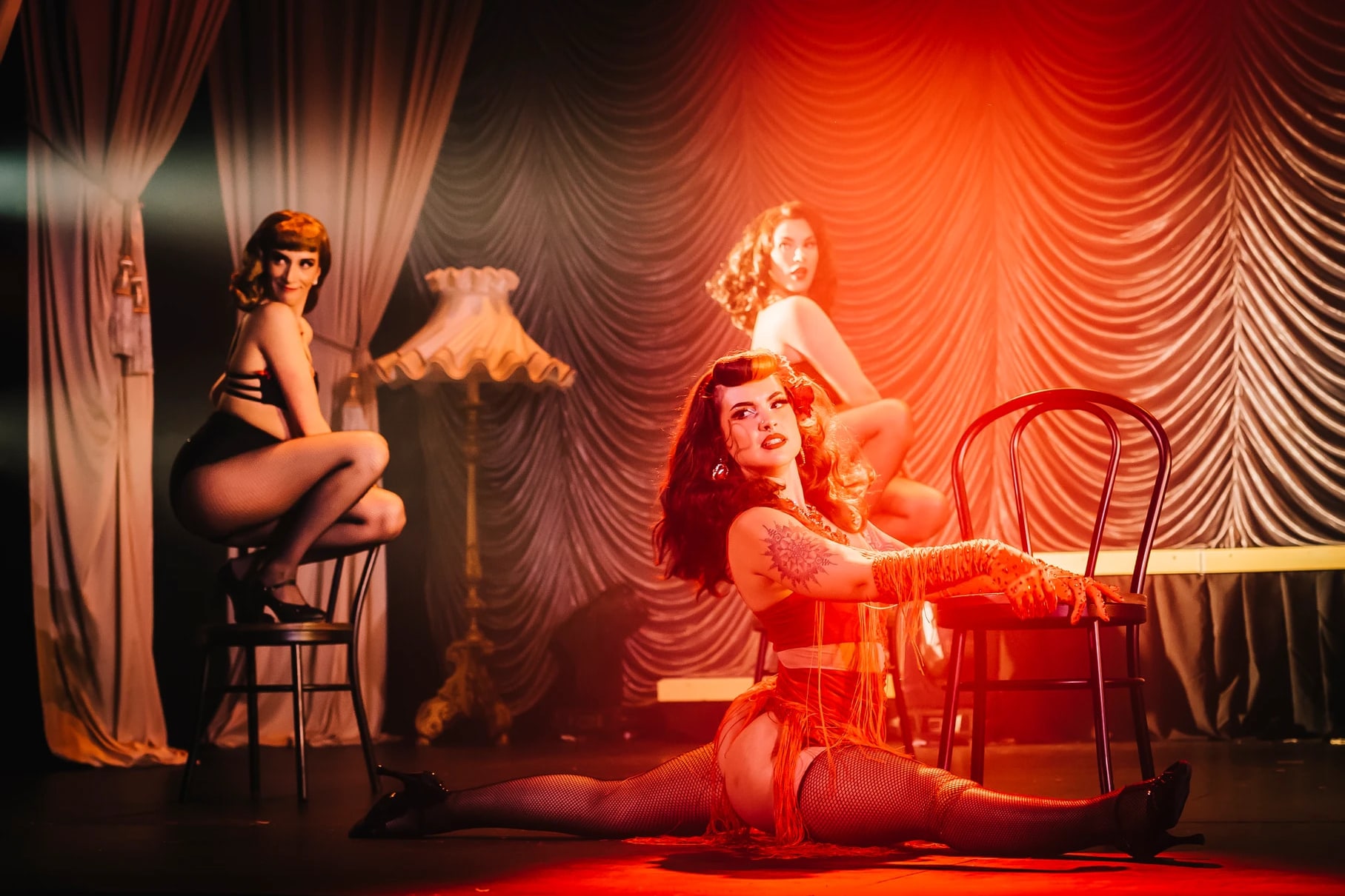 A burlesque performer doing the splits beside a chair under warm orange stage light