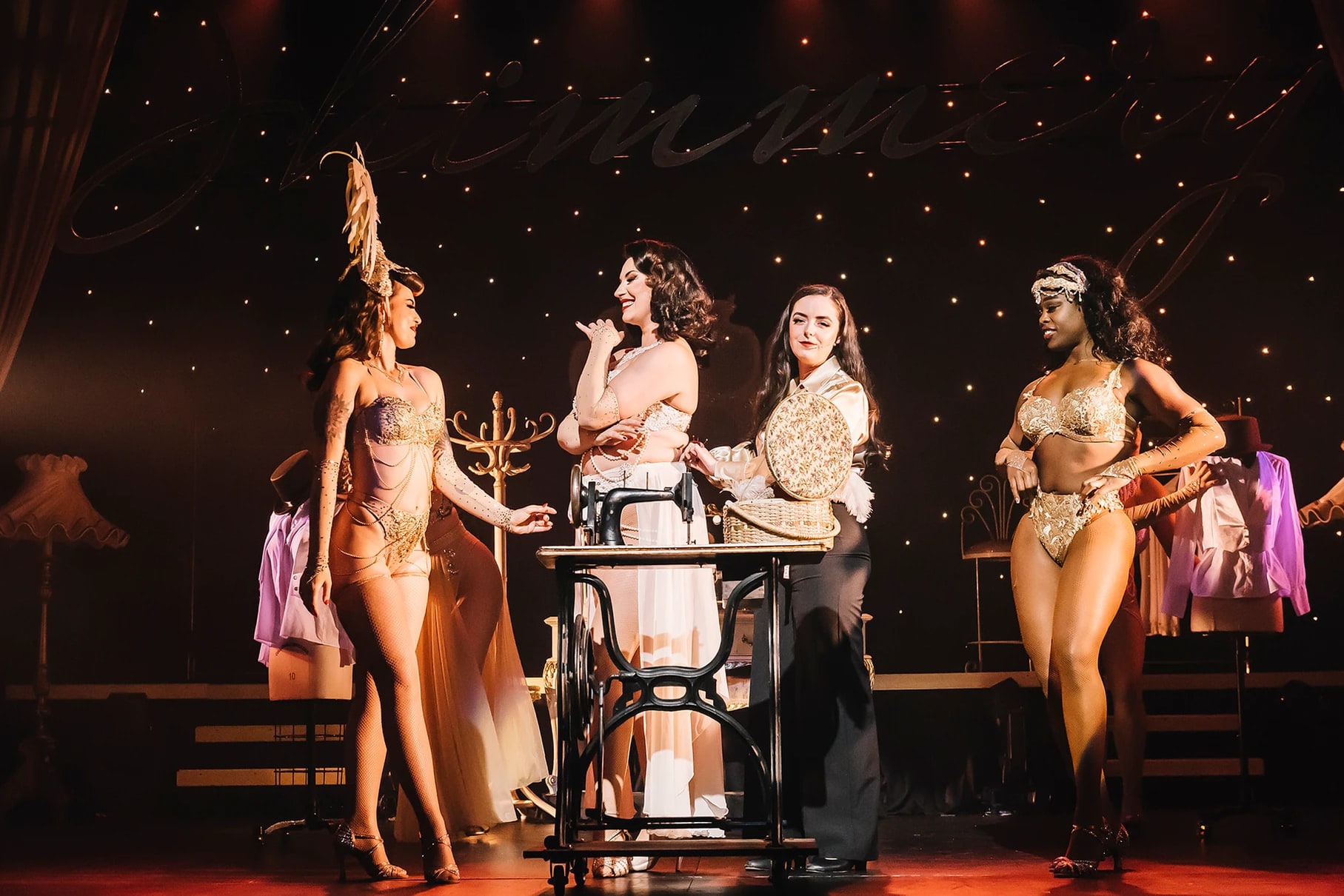 Lady Shimmery at her sewing machine surrounded by showgirls in rhinestone costumes