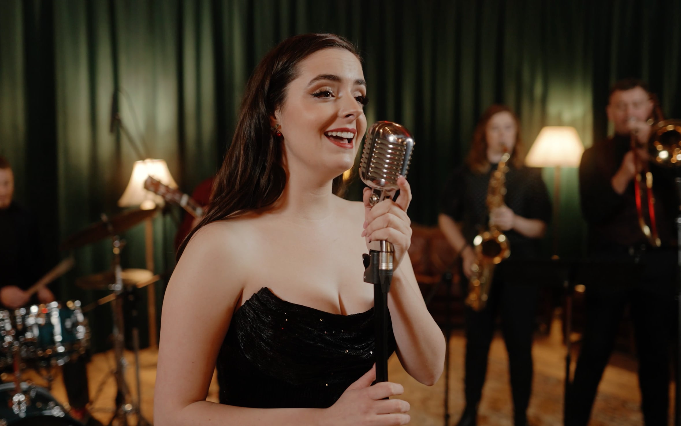 Close-up of Abbey smiling at a vintage microphone in a black strapless dress, with the band behind her